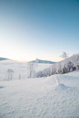 Morning idyll on the top of a snow-covered mountain with the rays of the sun irradiating the clouds, the ice cap of the mountain and a sliver of the moon. Beskydy mountains, Czech republic