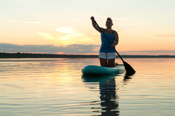 A woman on a kneeling SUP board with an oar at sunset against a golden sky floats in the water of the lake.