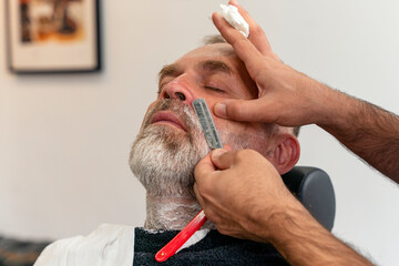 Barber trimming and cutting bearded man with shaving machine in barbershop. Hairstyling process.