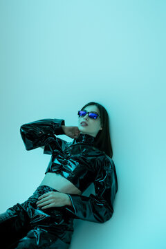 stylish young woman in sunglasses and black latex clothing sitting on blue background.