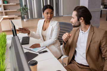 multiracial businesswoman pointing at computer monitor and talking to colleague in office.