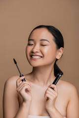 Fototapeta premium Cheerful asian woman with naked shoulders holding mascara isolated on brown.