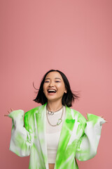 cheerful and stylish asian woman in fashionable jacket and necklaces showing wow gesture isolated...