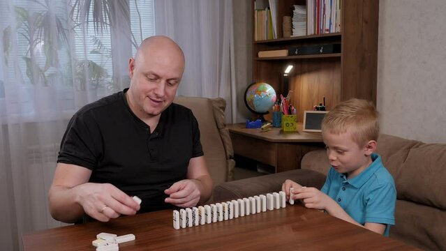 Cheerful father and son play with domino bones sitting at a wooden table, they put dominoes in a row.