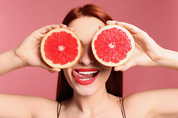 Beautiful redhead woman covering eyes with grapefruits on pink background