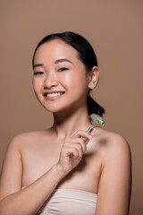 Positive asian woman in top holding jade roller and looking at camera isolated on brown.