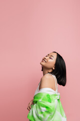 pleased asian woman with closed eyes posing with green and white jacket isolated on pink.