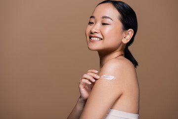 Cheerful asian model applying cosmetic cream on shoulder isolated on brown.