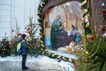 boy in winter clothes stands in front of a den with the holy family, the virgin mary, jesus, prays....
