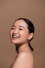 Positive asian model with naked shoulder looking at camera isolated on brown.