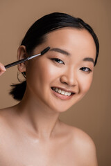 Obraz na płótnie Canvas Smiling asian model holding eyebrow brush and looking at camera isolated on brown.