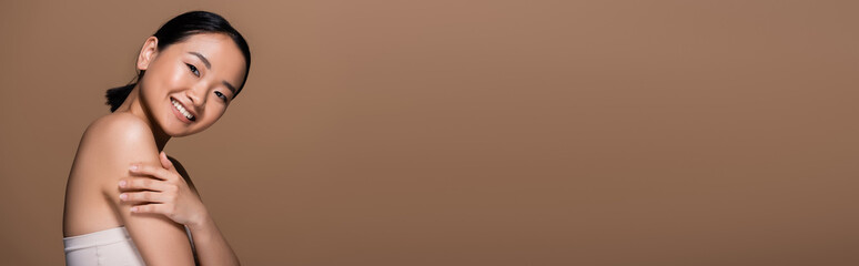 Cheerful asian woman in top touching arm and looking at camera isolated on brown, banner.