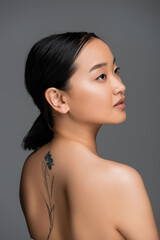 Brunette asian woman with tattoo on back looking away isolated on grey.