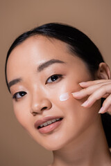 Portrait of young asian woman applying face cream on cheek isolated on brown.