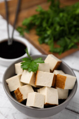 Bowl of smoked tofu cubes, soy sauce and parsley on white wooden table, closeup