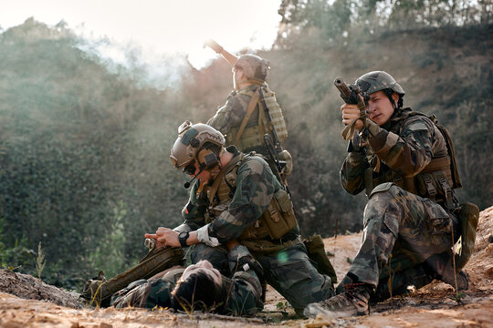brave european caucasian soldier help injured comrade lying on ground, in mountains, outdoors, another soldier calls for halp in the background. Fully equipped military forces in operations.