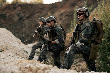 Fototapeta premium Group of armed special forces soldiers with rifles standing together looking at side, planning strategy of shooting, waiting for enemy. confident serious soldiers in military clothes outdoors