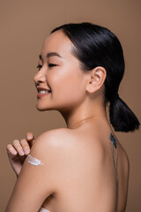 Cheerful asian model with naked shoulders applying cosmetic cream and looking away isolated on brown.