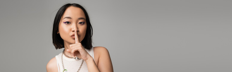 Fototapeta premium young asian woman with makeup and blue eyeliner showing hush sign isolated on grey, banner.