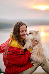 woman enjoying the awesome view of nature landscape, sitting on the top of mountain during summer vacation, travelling with pet spitz dog, dressed in colorful sportive outfit, river in background