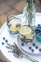 Two glasses with cool delicious homemade lemonade made of fresh blueberries, lemon and lavender on white round table on backyard in summer day.