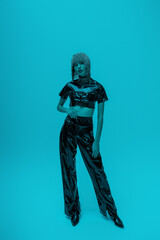 Full length of stylish model in headwear and latex clothes posing on blue background with lighting.