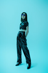 full length of stylish young woman in latex crop top and trousers standing on blue.