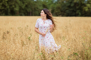 Fototapeta na wymiar A beautiful brunette woman in a white dress runs along a field of golden chickpeas ear. Stylish girl in the field in windy weather