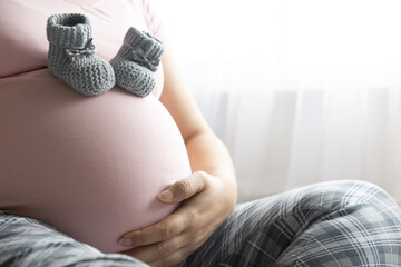 Pregnant woman in pink t-shirt touching her belly. Two little wool booties on belly. Healthy pregnancy.