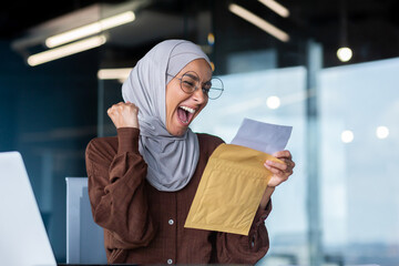 Successful businesswoman in hijab celebrating victory and successful achievement muslim woman inside office with laptop received good results notification mail female worker holding hand up triumph.