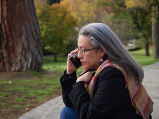 Older woman with gray hair and glasses wearing and talking on a mobile phone. Sitting on stone stairs in a park with a green grass background
