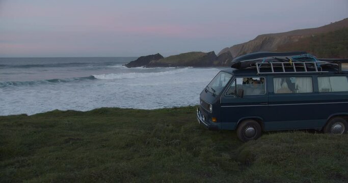 Camper van parking at the cliffs in a lonely bay during blue hour after sunset while big waves are rolling in the background in Galicia, Spain