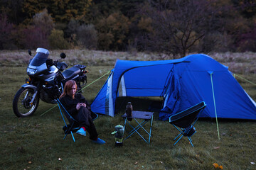 Night camp on the wilderness. Camping with big blue tent and touring motorcycle, Woman is sitting on folding chairs use smatphone. Concept of active lifestyle and tourism equipment