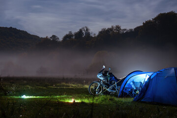 Camping, blue tent with light, touring motorcycle for travel. The concept of recreation and tourism. Space for text, selective focus. Mist, fog and moonlight.