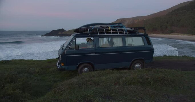 Camper van parking at the cliffs in a lonely bay during blue hour after sunset while big waves are rolling in the background in Galicia, Spain