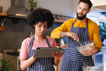 Biracial young woman watching recipe on digital tablet while boyfriend mixing batter in bowl at home