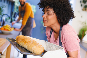 Biracial woman with eyes closed smelling freshly baked loaf of bread while cooking with boyfriend