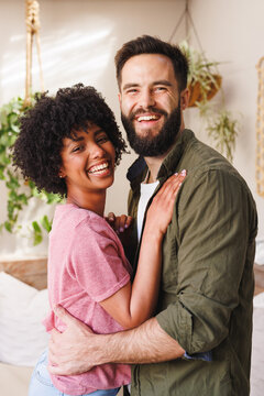 Portrait of cheerful biracial young couple laughing and hugging while standing against white wall