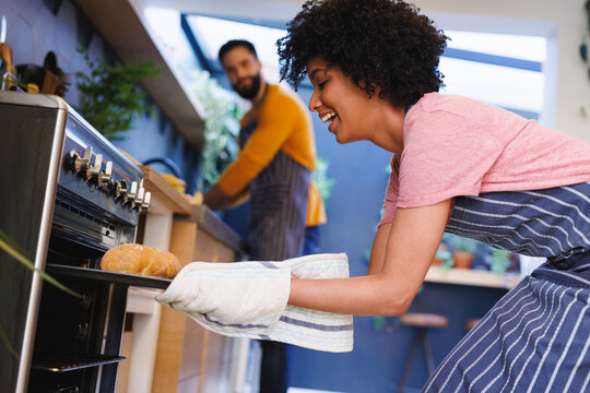 Biracial young man looking at cheerful girlfriend removing freshly baked bread from oven in kitchen