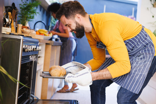 Biracial bearded man removing freshly baked loaf of bread from oven while cooking with girlfriend