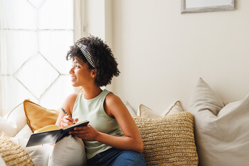 Thoughtful biracial young woman with afro hair looking through window while writing in diary on sofa
