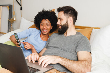 Smiling biracial young woman showing smartphone to boyfriend using laptop while relaxing on bed