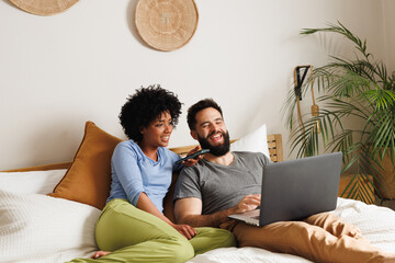 Cheerful biracial young couple watching moving over laptop while relaxing on bed against wall