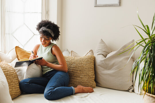 Smiling biracial young woman with afro hair writing in diary while sitting by window on sofa
