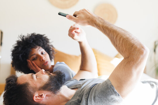 Biracial young woman looking at bearded boyfriend using smart phone while lying on bed at home