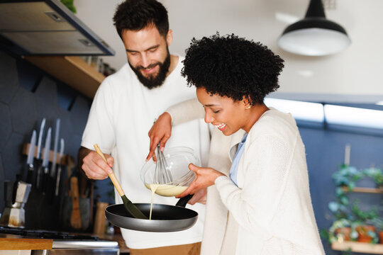 Happy biracial young couple pouring batter in cooking pan while making breakfast together in kitchen
