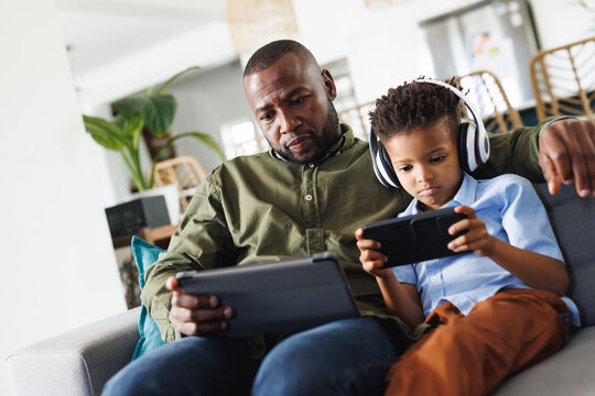 Happy african american father and son sitting on sofa, using tablet and smartphone in living room