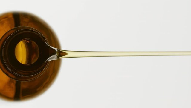 Vertical macro shot of brown reagent bottle is being tilted and yellow oil pours from it on grey background | Abstract anti-wrinkle cosmetics ingredients formulating concept