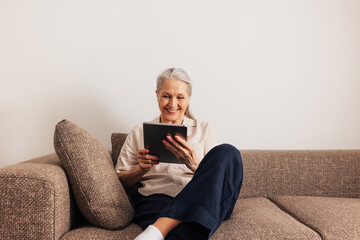 Obraz premium Adult cheerful female sitting on a sofa with digital tablet. Smiling senior woman holding a portable computer in the living room.