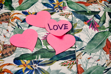 Four pink heart-shaped Post it on one of them written the word love, on top of a table with a tropical pattern.